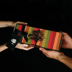 A rectangular gift box with a colorful striped pattern in red, orange, green, and yellow, featuring a black silhouette of a tree on top. The box is held by multiple hands against a solid black background, with a black ribbon and tag attached.