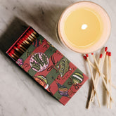 A decorative matchbox filled with red-tipped matches sits on a light marble surface beside a lit candle and loose matches.