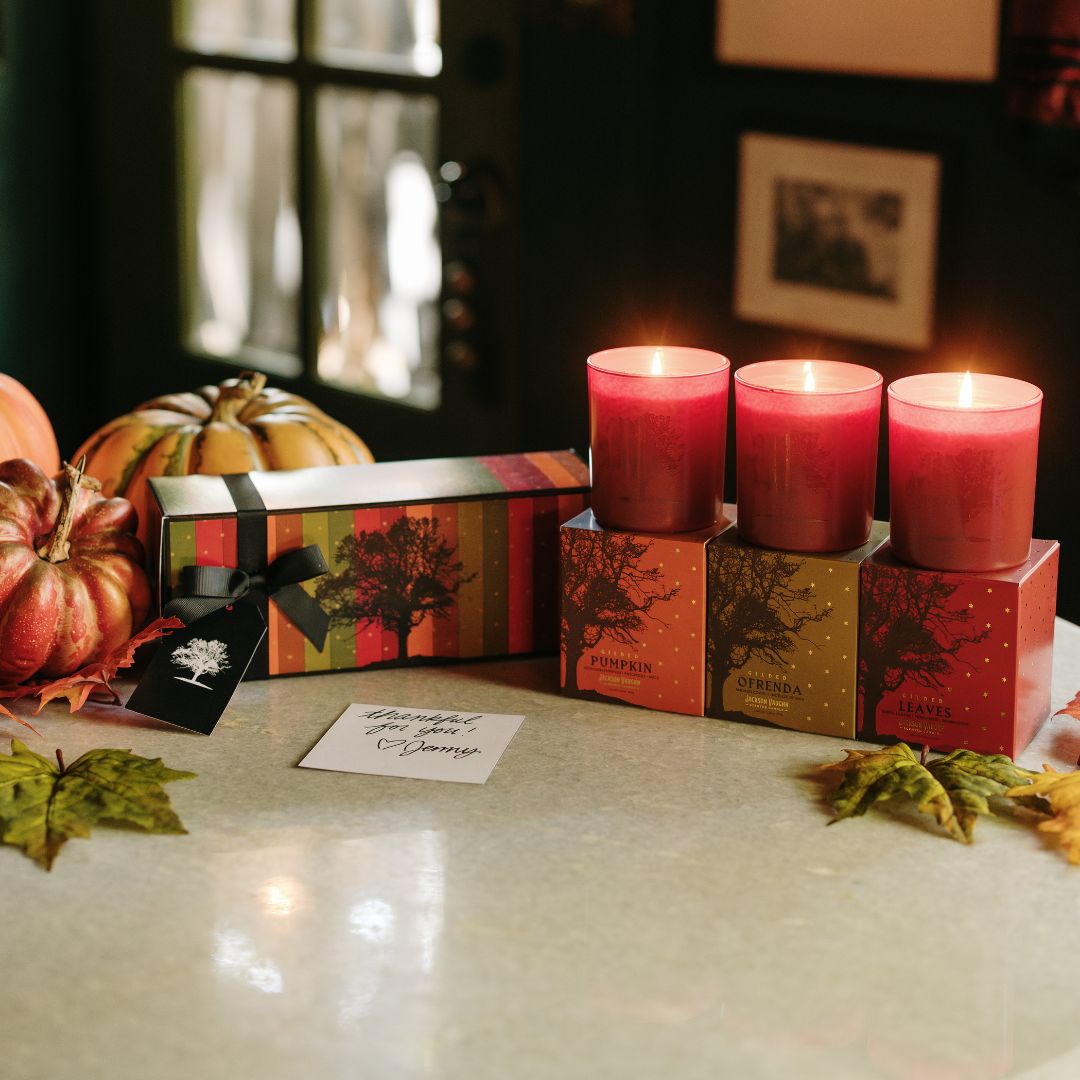 Three lit red pillar candles displayed on decorative boxes next to a matching gift box with a black ribbon, set on a tabletop with pumpkins, autumn leaves, and a handwritten note in a dimly lit indoor setting.