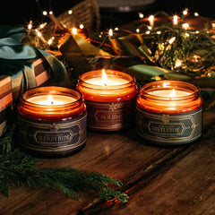 Three lit holiday candles in amber glass jars with decorative labels, arranged on a wooden surface with evergreen branches, a wrapped gift, and warm string lights in the background.
